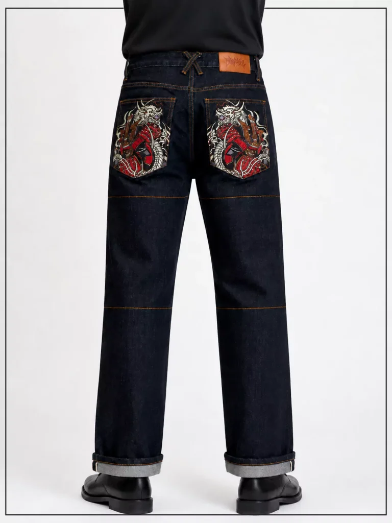Back view of dark denim jeans with ornate dragon embroidery on both rear pockets, orange contrast stitching, leather patch at waistband, cuffs rolled up, model wearing black top.